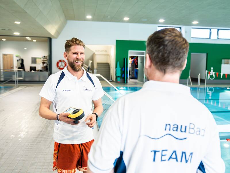 Zwei Männer in Sportbekleidung stehen lachend an einem Hallenbad, wobei einer ein technisches Gerät in der Hand hält.
