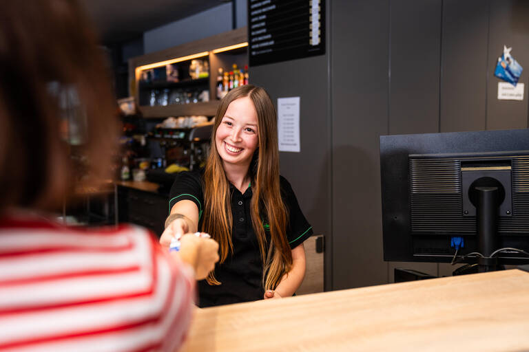 Eine lächelnde Frau hinter einem Tresen in einem Café reicht einer Person ein Ticket.