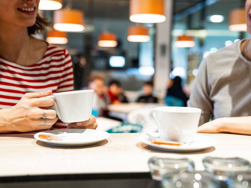 Zwei Personen sitzen an einer Bistrobar und trinken Kaffee. Im Hintergrund ein erleuchtetes Bistro mit orangene Lampen und Gästen