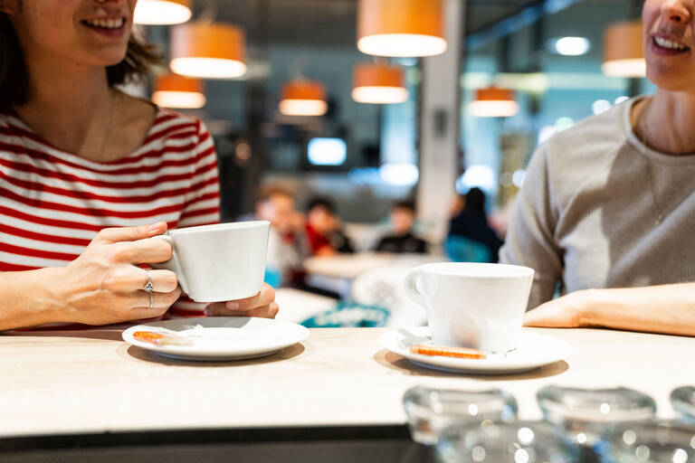 Zwei Personen sitzen an einer Bistrobar und trinken Kaffee. Im Hintergrund ein erleuchtetes Bistro mit orangene Lampen und Gästen
