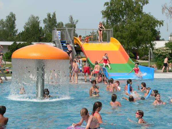 Mehrere Kinder spielen und planschen in einem Schwimmbecken mit einer bunten Rutsche und einem pilzförmigen Wassersprinkler.