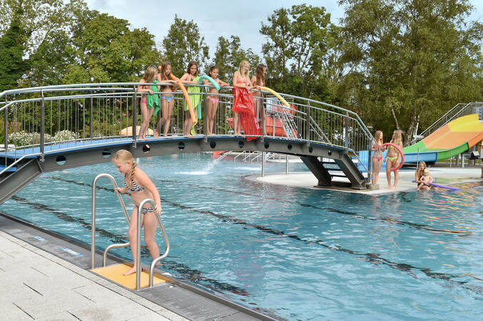 Kinder in Badekleidung stehen auf einer Brücke über einem Außenpool, während ein Mädchen die Pooltreppe hinuntersteigt.