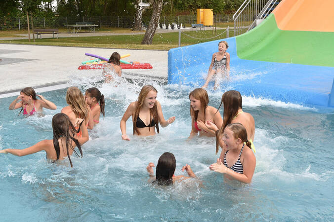 Eine Gruppe von Kindern spielt fröhlich in einem Pool mit einer Wasserrutsche im Hintergrund.