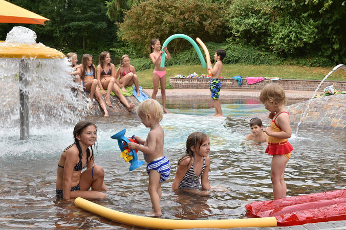 Mehrere Kinder spielen in einem flachen, wasserreichen Bereich mit Fontänen, wobei einige am Rand sitzen und andere im Wasser Planschspielzeug benutzen.