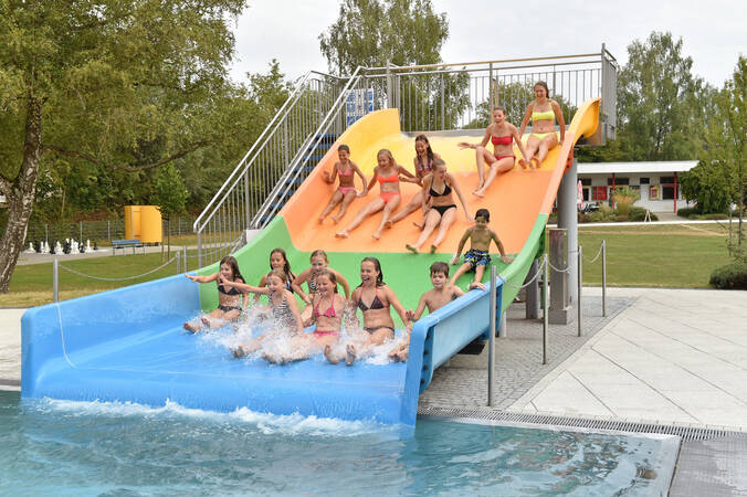 Mehrere Kinder rutschen lachend eine dreifarbige Wasserrutsche hinunter in ein Schwimmbecken.