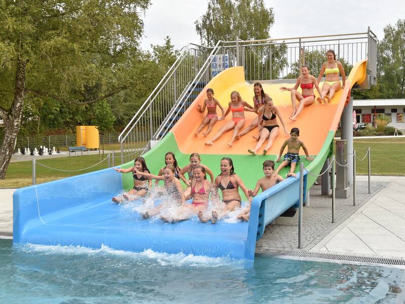 Mehrere Kinder rutschen lachend eine dreifarbige Wasserrutsche hinunter in ein Schwimmbecken.