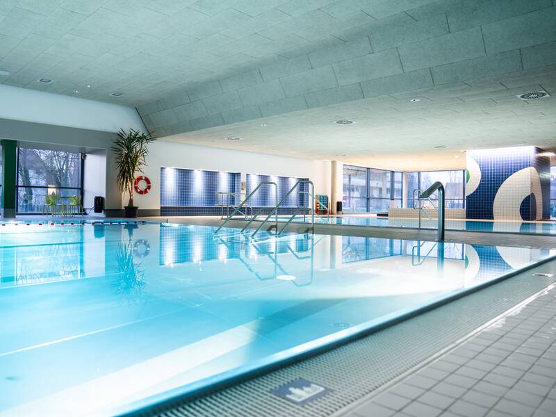 Ein modernes, gut beleuchtetes Hallenbad mit einem großen Pool, an dem Pflanzen und bunte Wandfliesen sichtbar sind.