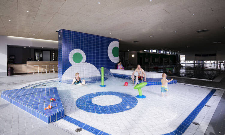 In einem Kinderplanschbecken des Hallenbads Naubad spielen mehrere Kinder im flachen Wasser umgeben von blauen Fliesen und Wasserspielzeugen.