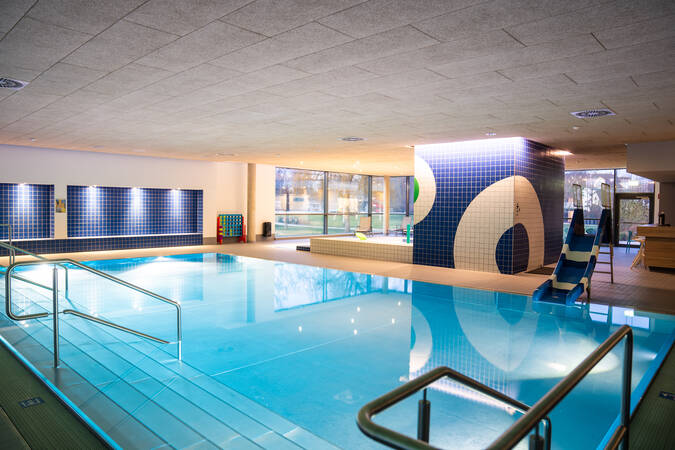 Ein modernes, gut beleuchtetes Hallenbad mit einem Pool für Nichtschwimmer, an dem Pflanzen und bunte Wandfliesen sichtbar sind.