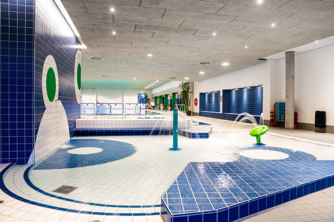 Das Bild zeigt ein modernes Hallenbad mit blauen und grünen Fliesen, einem flachen Kinderbecken und Wasserspielen unter einer hellen Decke.