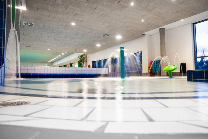 Das Bild zeigt ein modernes Hallenbad mit blauen und grünen Fliesen, einem flachen Kinderbecken und Wasserspielen unter einer hellen Decke.