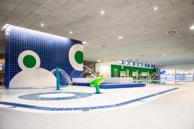 Das Bild zeigt ein modernes Hallenbad mit blauen und grünen Fliesen, einem flachen Kinderbecken und Wasserspielen unter einer hellen Decke.