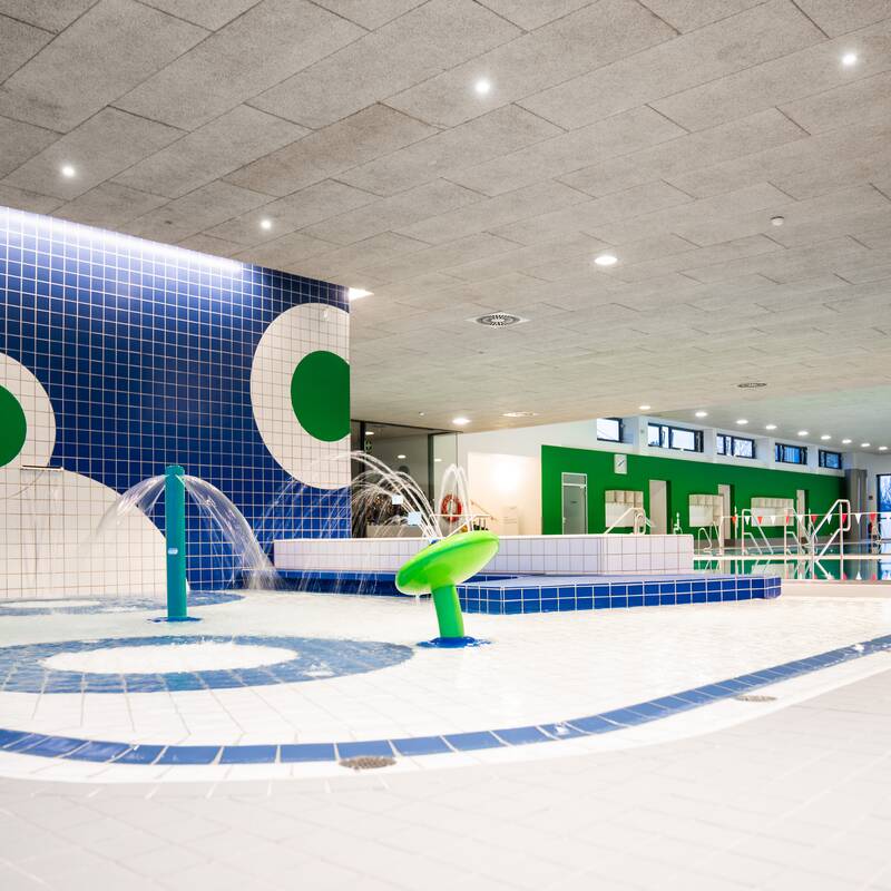 Das Bild zeigt ein modernes Hallenbad mit blauen und grünen Fliesen, einem flachen Kinderbecken und Wasserspielen unter einer hellen Decke.