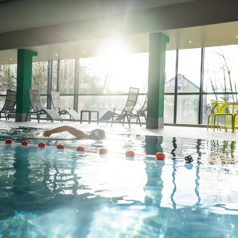 In einem modernen Hallenbad mit großer Fensterfront durch durch die die Sonne hereinscheint zieht ein Mann seine Bahnen in einem großen Schwimmbecken
