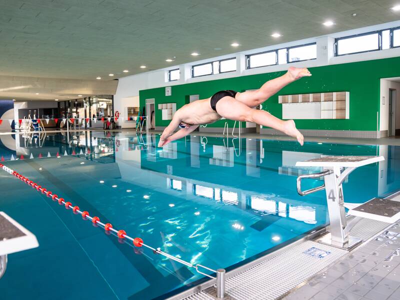 Eine Person in Badebekleidung springt von einem Sprungbrett ins Hallenbad.