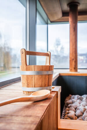 Ein hölzerner Wasserbehälter und eine Schöpfkelle stehen auf einer Holzbank neben einem Saunaofen mit Steinen in einem Raum mit großen Fenstern.