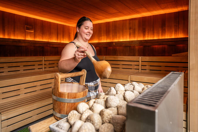Eine Person gießt mit einer Holzkelle Wasser aus einem Eimer über heiße Steine in einer holzverkleideten Sauna.