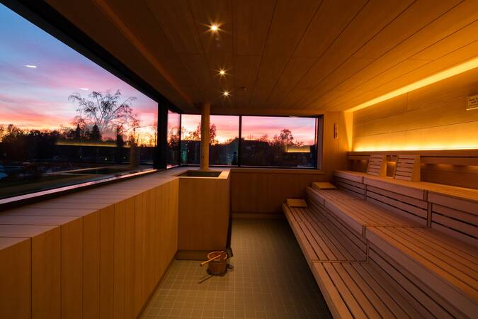 Eine moderne Sauna mit Holzinterieur, mehreren Sitzreihen und einem großen Fenster, das Abendstimmung hereinlässt.