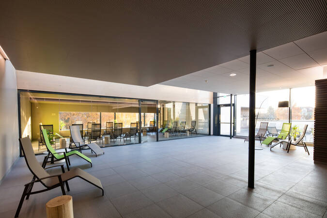 Der Raum zeigt eine entspannende Lounge mit mehreren Liegestühlen, großen Glasfenstern und Blick auf einen Außenbereich.