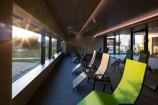 Der Raum zeigt eine entspannende Lounge mit mehreren Liegestühlen, großen Glasfenstern und Blick auf einen Außenbereich.
