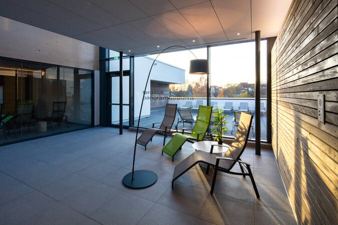 Der Raum zeigt eine entspannende Lounge mit mehreren Liegestühlen, großen Glasfenstern und Blick auf einen Außenbereich.