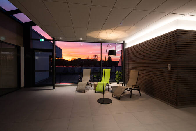 Der Raum zeigt eine entspannende Lounge mit mehreren Liegestühlen, großen Glasfenstern und Blick auf einen Außenbereich bei Abendstimmung