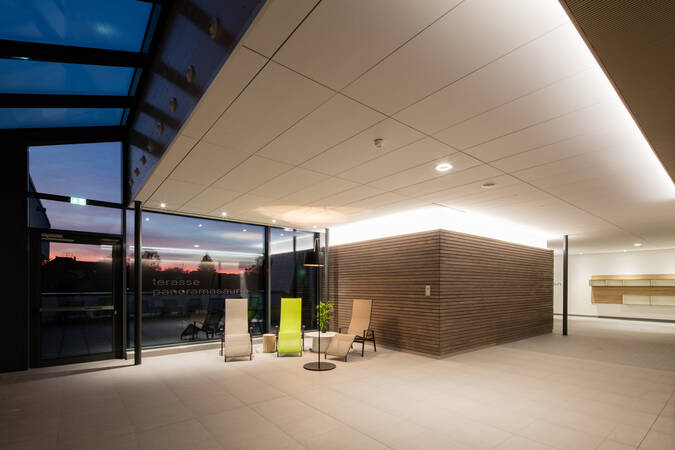Der Raum zeigt eine entspannende Lounge mit mehreren Liegestühlen, großen Glasfenstern und Blick auf einen Außenbereich bei Abendstimmung