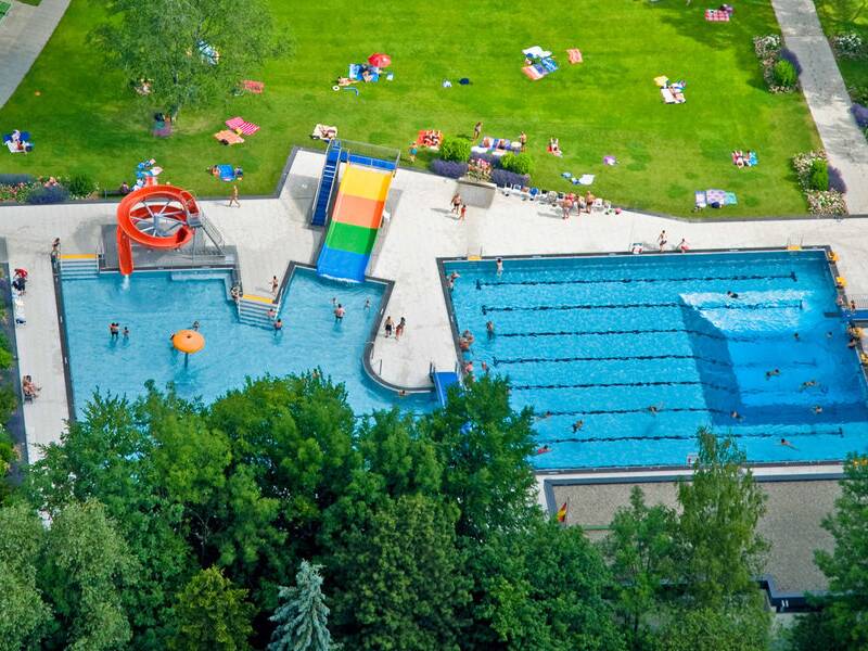 Eine Luftaufnahme zeigt das Freibad Naubad mit zwei großen Schwimmbecken, einer Rutsche und einer grünen Liegewiese, auf der Menschen entspannen.