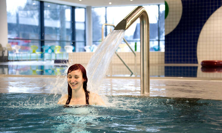 Eine Frau steht lächelnd im Wasser eines Hallenbads, während ein Wasserstrahl aus einer Metallröhre auf ihren Kopf fällt.