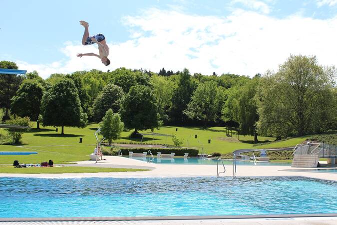 Ein Mann springt in einer saltierenden Bewegung von einem Sprungbrett in ein Schwimmbecken, umgeben von einer grünen, parkartigen Landschaft.